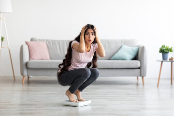 Shocked young Indian lady sitting on scales, terrified of weight gain at home, free space. Healthy...
