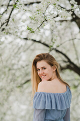 Young pretty caucasian blonde woman with freckles wearing natural makeup in light blue dress near the beautiful blooming spring tree by the lake. Youth, freshness, beauty, happiness, emotions concept.