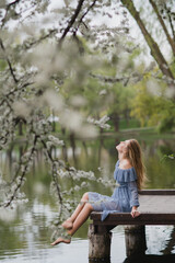 Young pretty caucasian blonde woman with freckles wearing natural makeup in light blue dress near the beautiful blooming spring tree by the lake. Youth, freshness, beauty, happiness, emotions concept.