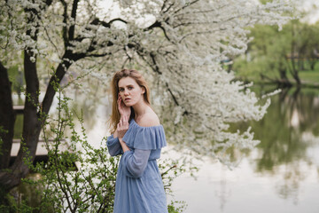 Young pretty caucasian blonde woman with freckles wearing natural makeup in light blue dress near the beautiful blooming spring tree by the lake. Youth, freshness, beauty, happiness, emotions concept.