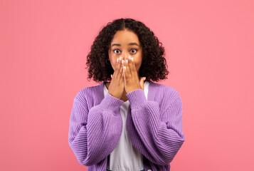 Shocked young black woman covering her mouth with hands and looking at camera over pink studio...
