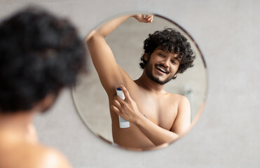 Shirtless indian man putting deodorant underarm after shower, looking at round mirror and smiling...