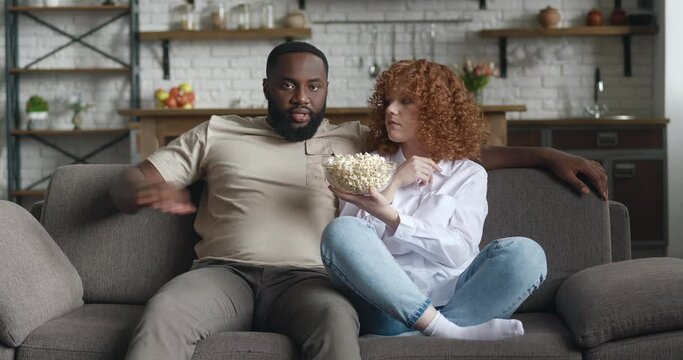 Young interracial family watching comedy movie on TV with popcorn sitting on couch in apartment. Laughing couple watching funny video together on tv at home