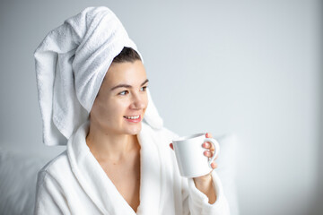 a young woman   after shower in white bathrobe and towel on head  sits on a bed in the morning at home  and drinks coffee. 