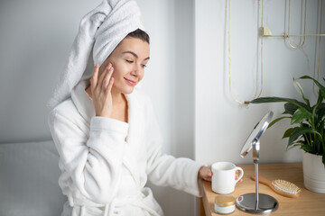 pretty woman  in white bathrobe and towel on head, putting cream on face skin, massaging face tenderly, having pleasure, enjoying morning time, self care concept - obrazy, fototapety, plakaty