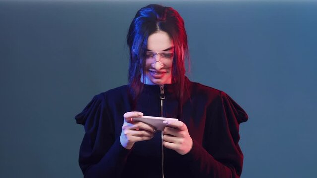 Phone gaming. Smartphone nerd. Cybersport gamer. Happy enthusiastic woman in red neon light playing cellphone app on blue background.