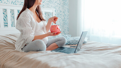 Pragnant woman puts coins into piggy bank. Pregnant with laptop on bed. Pregnant woman online shopping.