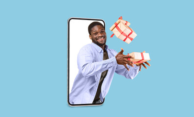 Smiling young attractive black guy hold gifts boxes on blank screen of big phone isolated on blue background