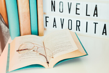 Medium close-up. Light box with message for the international book day, next to it covered books and an open book with a pair of glasses, on the bedside table in the bedroom.