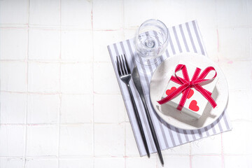  Valentine day table setting with plate, gift box with festive red ribbon, wine glass, fork and knife, red roses flowers bouquet white tiled table flatlay copy space