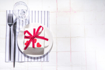  Valentine day table setting with plate, gift box with festive red ribbon, wine glass, fork and knife, red roses flowers bouquet white tiled table flatlay copy space