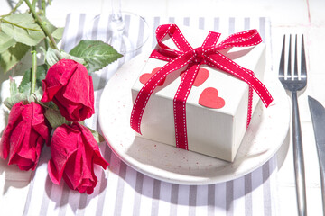  Valentine day table setting with plate, gift box with festive red ribbon, wine glass, fork and knife, red roses flowers bouquet white tiled table flatlay copy space