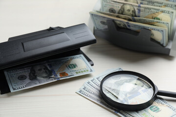 Modern currency detector with dollar banknotes and magnifying glass on white wooden table. Money examination device