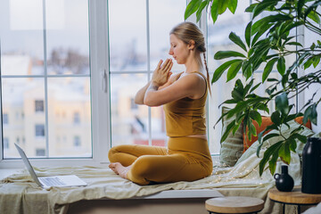 Young blonde long-haired woman in yellow meditates in lotus pose sitting on blanket covering windowsill and looking at laptop screen