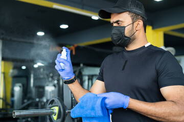 Young man with medical face mask and hand gloves sanitizing gym due to covid-19 infection - concept...