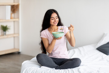 Full length portrait of beautiful millennial Indian lady having cereal with milk while sitting on...