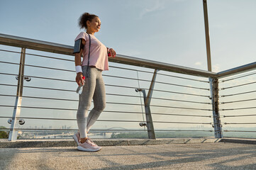 Full length portrait of a sportswoman, female athlete with a skipping rope, standing on the modern glass city bridge, relaxing after cardio workout outdoor. Sport, fitness, active lifestyle concept