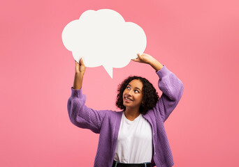 Positive black woman holding empty speech bubble above her head on pink studio background, mockup...
