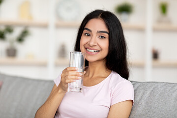 Smiling young Indian woman holding glass of fresh clean water while sitting on couch in living room. Keep hydrated concept