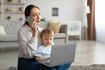Busy and stressed mother talking on cellphone while her toddler son sitting on mom's knees and looking at laptop
