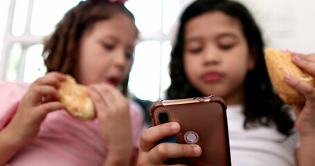 Generation Z little girls using mobile phone while eating sandwich snack