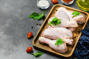 Raw chicken legs, parsley, olive oil, tomatoes on wooden tray on dark background. Top view, copy space.