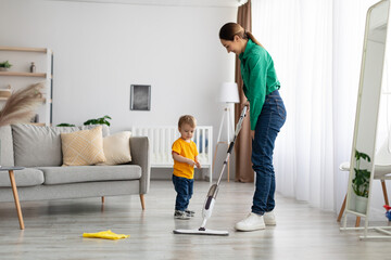 Young mother teaching toddler son cleaning at home, mopping the floor in living room interior, free space