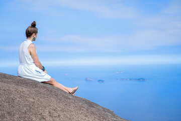 Fototapeta na wymiar A woman in a light dress is relaxing on a top of the rock against the backdrop of the ocean and sky on a sunny day in brazil
