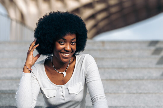 Portrait of a smiling young black woman sitting on a staircase in the street and with her hand stroking her afro hair