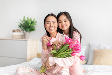 Obraz na płótnie Canvas Cheerful chinese young girl hug millennial lady in pink pajama with tulips bouquet on bed in bedroom interior