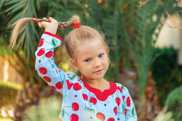 Portrait of a beautiful little girl who smiles and carelessly plays with her pigtail, holding her...