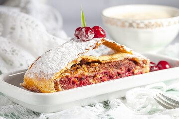 
A piece of freshly baked homemade strudel with cherries and powdered sugar, on a rectangular white porcelain plate on a light background