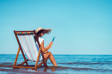 Back view of woman in sunhat who using laptop while lying on the beach chaise longue at the seaside - obrazy, fototapety, plakaty