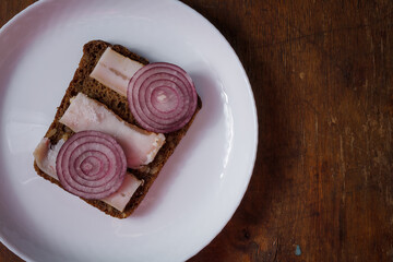 whole grain rye bread and prickled pork lard sandwich with red onions on white plate, wooden table. Ukrainian cuisine appetizer, close up shot