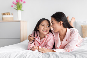 Smiling pretty cute korean millennial woman hugging teenage daughter in pink pajama lie on bed in...