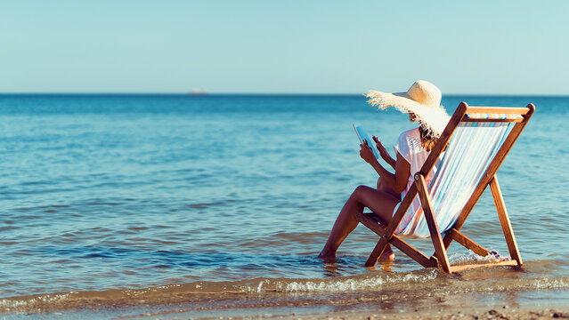 Rear view girl in sunhat working at laptop while lying on the beach chaise longue at the seaside