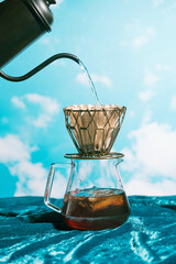Water pouring into metall dripper with paper filter and iced coffee in front of blue sky, alternative brewing