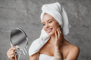 Beauty rituals. Happy lady with bath towel on head touching her glowing skin and looking in...