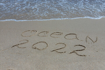 The inscription ocean 2022 on the sand by the water and the rising wave, seashore beach vacation by the sea