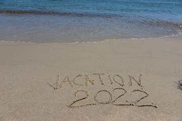 The inscription vacation 2022 on the sand by the water and the rising wave, seashore beach vacation by the sea