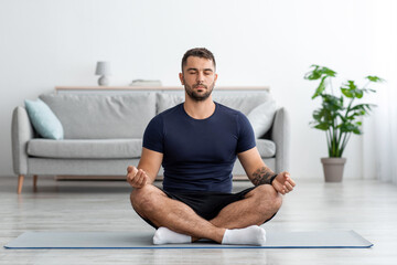 Serious calm concentrated young caucasian muscular man with closed eyes sits on floor in lotus position, practices yoga