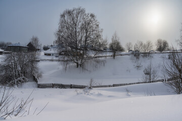 A rural winter landscape with snow-covered houses and gardens on a hill, a mighty tree and northern sun. Cherdyn (Ural, Russia)