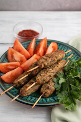 Blurred image of lula kebabs in a plate with tomato slices and greens on a light background.