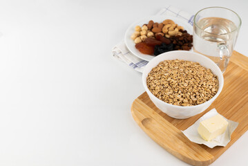 On a white background, oatmeal in a plate, a piece of butter, a glass of water on a board and a plate with dried fruits and nuts.