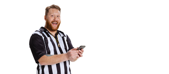 Excited young bearded man, soccer or football referee using phone isolated on white studio...