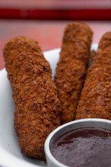 Finger food. Closeup view of fried breaded mozzarella sticks with dipping barbecue sauce in a white bowl on the red wooden table.
