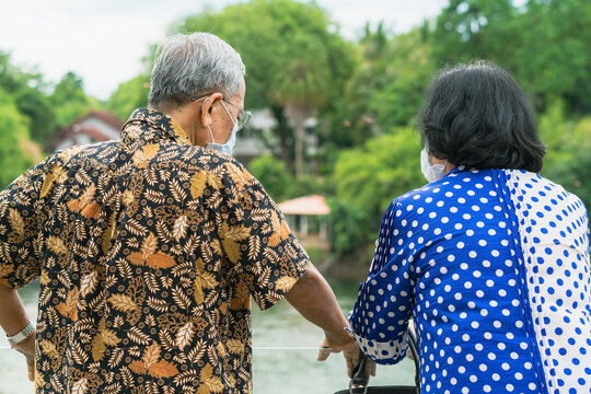 Back view of elderly couple wear protective face mask due to coronavirus (Covid-19) standing side by side at riverside. Relaxed lifestyle for two retired people enjoying freedom in summer holidays.