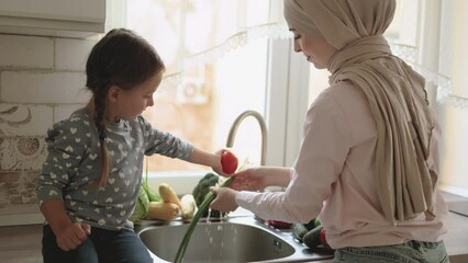 Beautiful muslim woman with her four year old daughter is washing vegetables in the kitchen sink. Fresh vegetables, onions, tomatoes, peppers, cucumbers, broccoli, cabbage. Healthy food for children