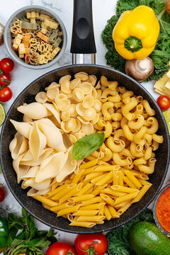 Frying pan with assorted raw pasta amidst vegetables