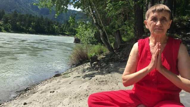 an elderly woman doing yoga on the banks of a mountain river, folded her hands in a namaste greeting, summer sunny day